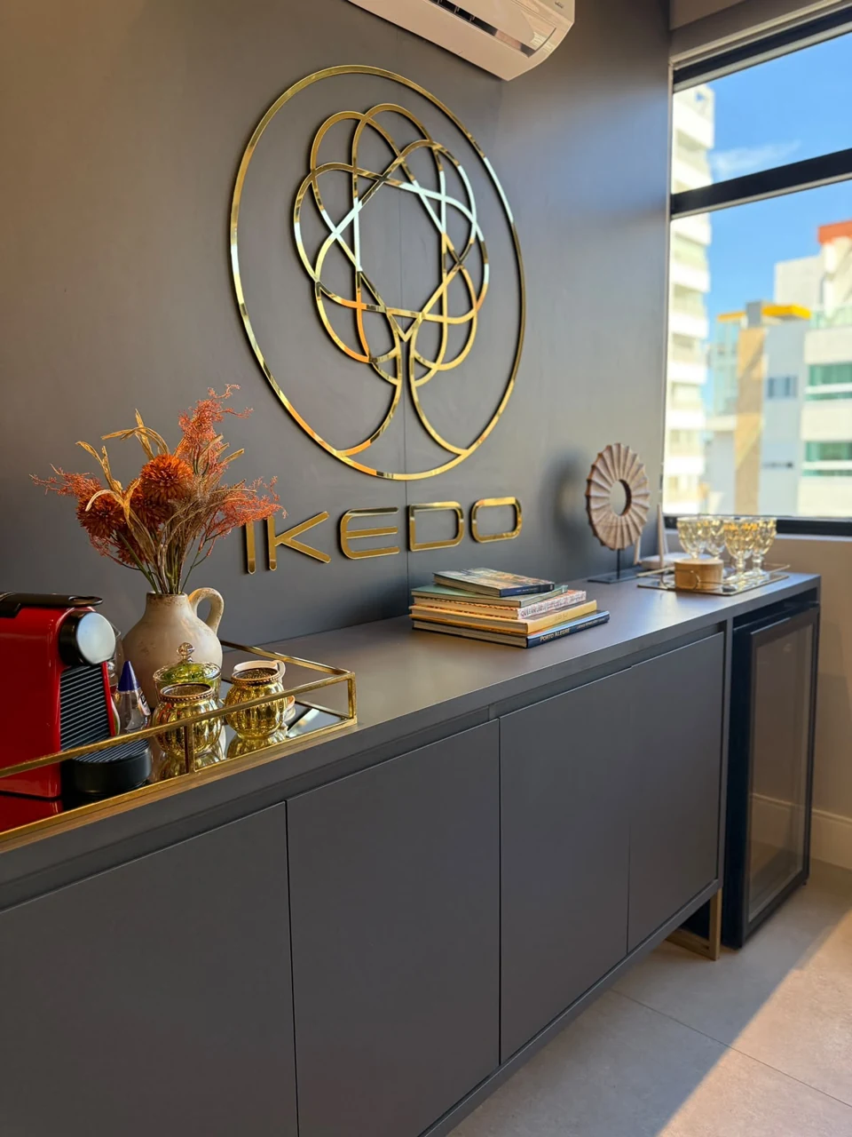 Logotipo dourado IKEDO na parede do escritório, ao lado de livros e objetos de acolhimento